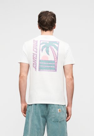 Személy fehér pólóban, amelyen pasztell színű pálmafás, naplementés grafika és a "Santa Cruz" felirat található a háton, kékkel tengerzöld rövidnadrággal párosítva.