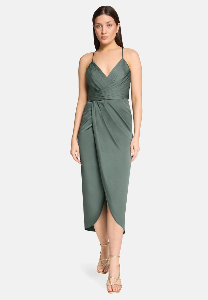 Femme portant une robe portefeuille verte sans manches avec des bretelles fines et des talons dorés à brides, debout devant un fond clair uni.