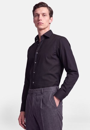 Jeune homme portant une chemise noire à manches longues boutonnée, rentrée dans un pantalon gris plissé, debout avec une main dans la poche.