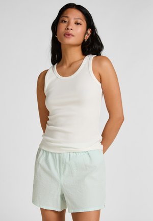 Vrouw met lang zwart haar, gekleed in een witte, mouwloze, geribbelde tanktop en lichtgroene, gestructureerde korte broek, poseert met haar handen in de zakken.