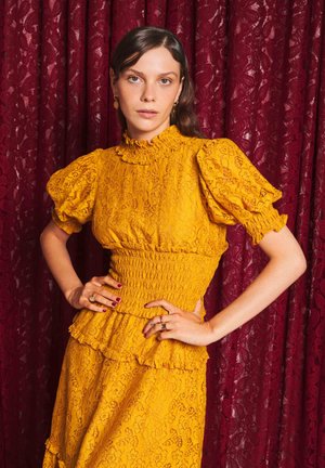 Femme portant une robe en dentelle jaune avec des manches bouffantes, debout les mains sur les hanches devant un rideau à motif rouge foncé.