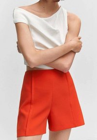 Person iført en hvid én-skulder crop top og højtaljede lyse orange shorts, med armene korslagt over brystet foran en ensfarvet baggrund.