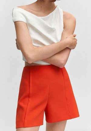 Persoon in een witte off-shoulder crop top en feloranje hoge shorts, met de armen over elkaar tegen een effen achtergrond.