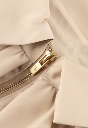 Gouden rits met MK-logo op beige stof, met gedetailleerde stiksels en samengeplooide stof rond het ritsgebied.