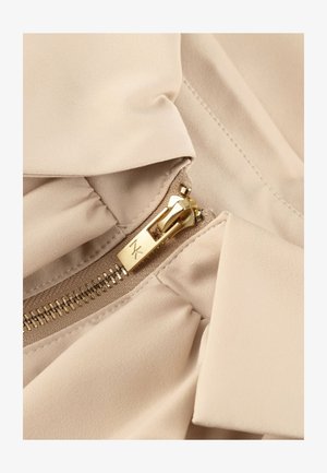 Gouden rits met MK-logo op beige stof, met gedetailleerde stiksels en samengeplooide stof rond het ritsgebied.