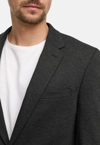Mørk grå blazer laget av teksturert stoff, med knappede reverser og en enkelt brystlomme. Kombinert med en hvit T-skjorte under.