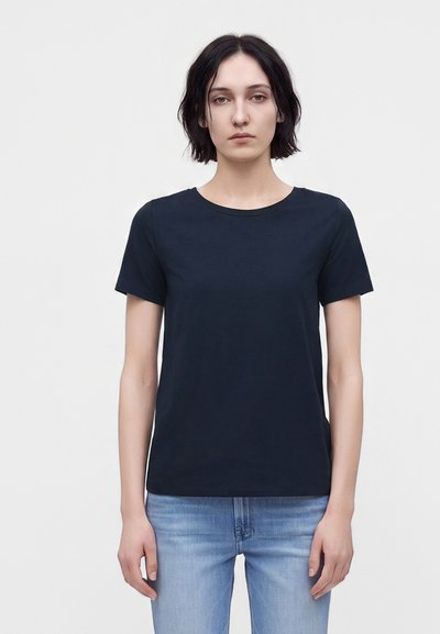 Mujer con cabello corto y oscuro, que lleva una camiseta negra lisa de manga corta y vaqueros azul claro, de pie sobre un fondo blanco.