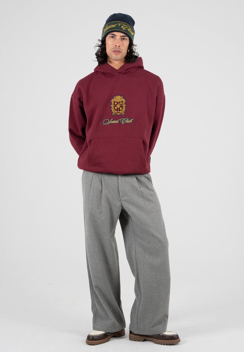 Burgundowa bluza z kapturem z haftowanym logo i tekstem, połączona z szarymi spodniami o wysokim stanie z szerokimi nogawkami oraz brązowymi butami z białymi akcentami.
