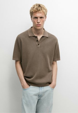 Junger Mann mit kurzen lockigen blonden Haaren, der ein braunes kurzärmeliges Strick-Poloshirt und hellblaue Jeans trägt, steht mit den Händen in den Taschen.