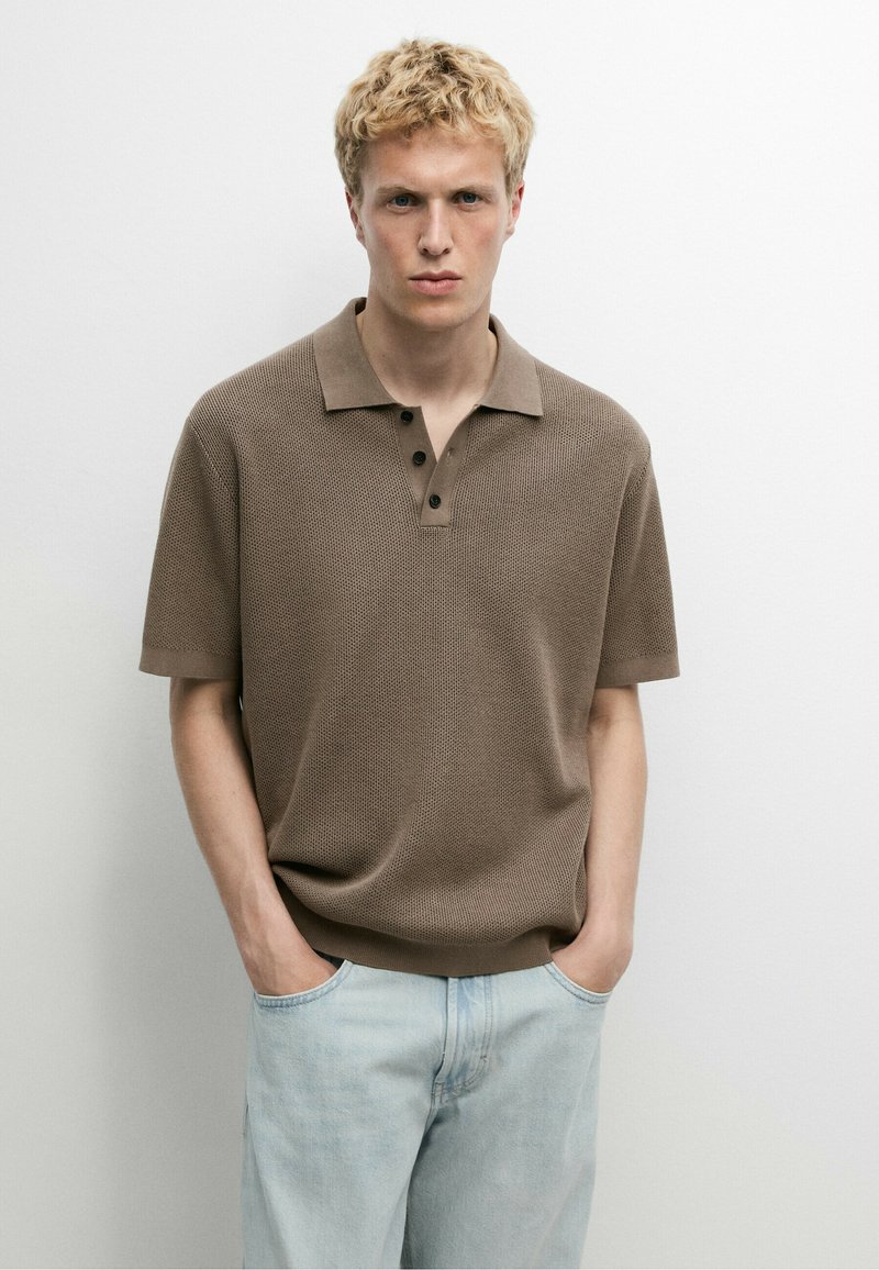 Jeune homme aux cheveux blonds courts et bouclés, portant un polo en maille marron à manches courtes et un jean bleu clair, debout les mains dans les poches.