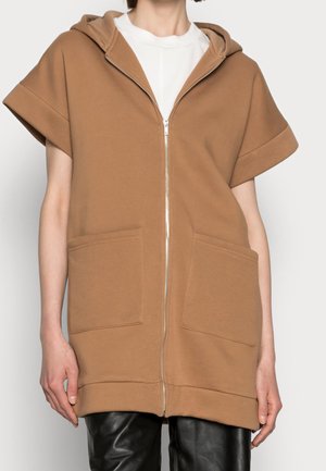 Femme portant un gilet zippé à capuche beige à manches courtes avec poches devant, par-dessus une chemise blanche et un pantalon noir.