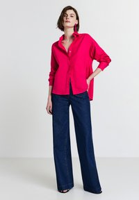 Donna che indossa una camicia oversize rosa brillante, orecchini abbinati, jeans a gamba larga scuri, in piedi con una mano in tasca su uno sfondo neutro.