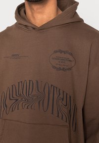 Felpa marrone con cappuccio, caratterizzata da un grande design di testo distorto "GOODFORNOTHING" in nero. Include un dettaglio del logo sulla parte superiore del petto.
