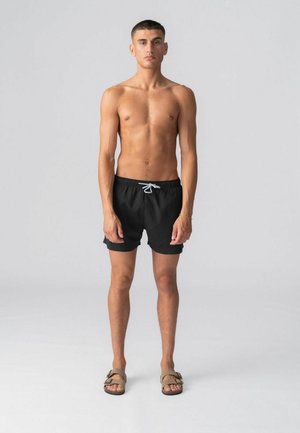 Młody mężczyzna stoi, bez koszulki, ubrany w czarne kąpielówki z białym sznurkiem i beżowe sandały, na jasnoszarym, jednolitym tle.