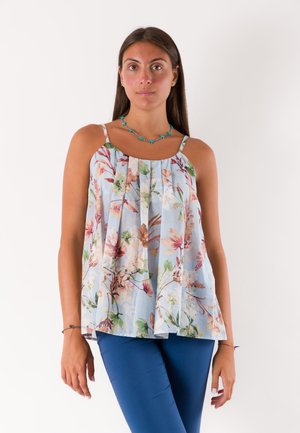 Donna che indossa un top senza maniche a motivo floreale e pantaloni blu, in piedi davanti a uno sfondo bianco uniforme con una collana turchese.