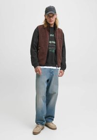 Brun fleecevest over en sort sweatshirt med grøn tekst, lyseblå denimjeans og beige sko. Afslappet look med en kasket.