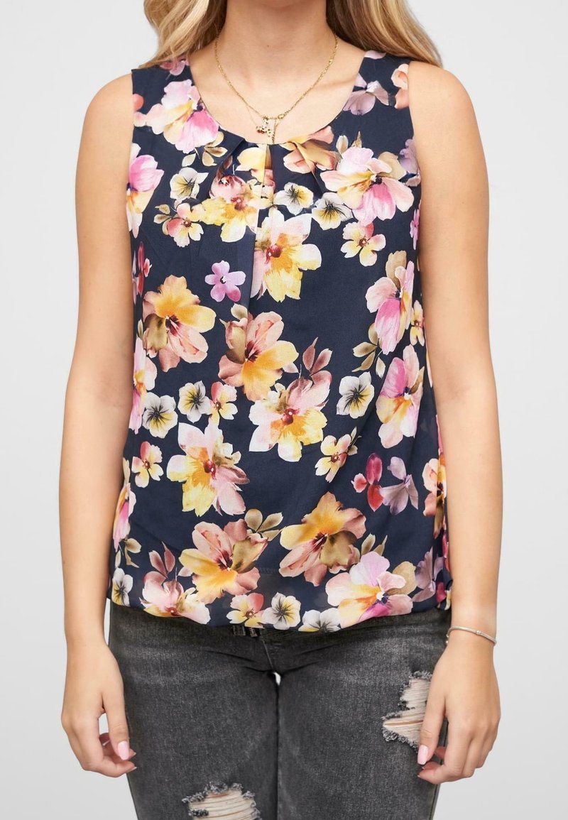 Femme portant un haut sans manches bleu marine avec un imprimé floral rose, jaune et blanc et un jean noir déchiré, visible du cou aux hanches.
