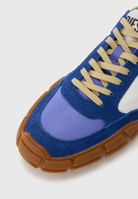 Blaue Wildleder-Sneaker mit lila Textil-Akzenten, beigen Schnürsenkeln und einer Gummisohle aus Kaugummi. Verfügt über ein geschwungenes Design und gestickte Details.