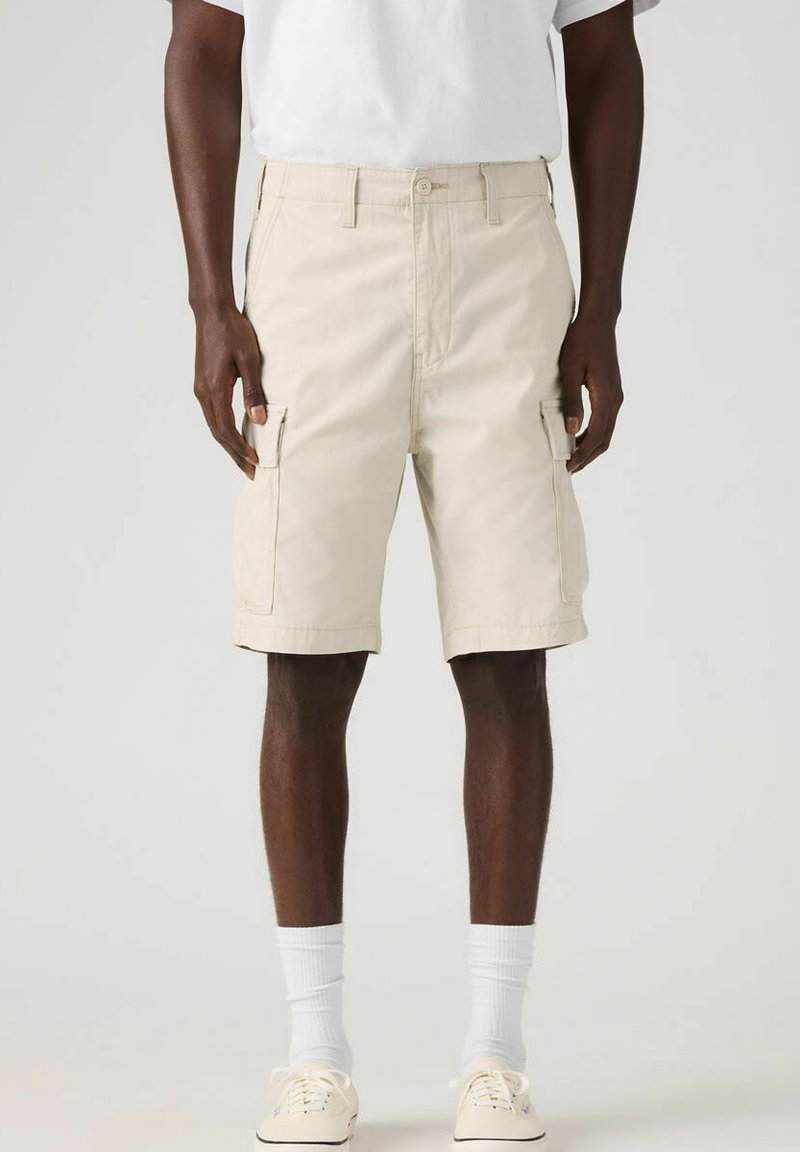 Un short cargo beige en coton, doté de plusieurs poches latérales, d'une coupe droite et d'un ourlet retroussé. Porté avec un t-shirt blanc uni et des baskets.