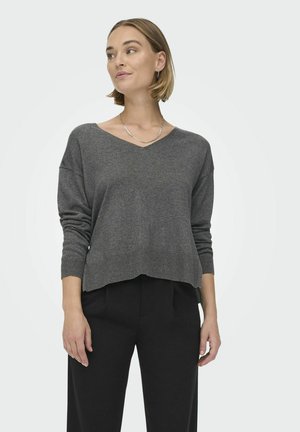 Grauer V-Ausschnitt-Pullover mit langen Ärmeln, lockerer Passform und geripptem Saum, kombiniert mit schwarzen Hosen. Strukturiertes Strickgewebe.