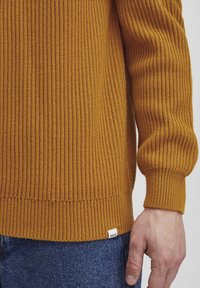 Mustard knitted sweater with ribbed texture, fitted cuffs, and a small brand tag on the lower hem. Paired with dark blue denim jeans.