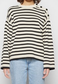 Pull rayé crème et noir, avec un col et des poignets côtelés. Deux boutons noirs accentuent l'épaule. Porté avec un jean bleu clair.