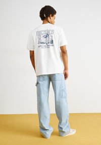 T-shirt en coton blanc avec un design graphique dans le dos, associé à un jean cargo bleu clair. La scène présente un fond uni et un sol jaune.