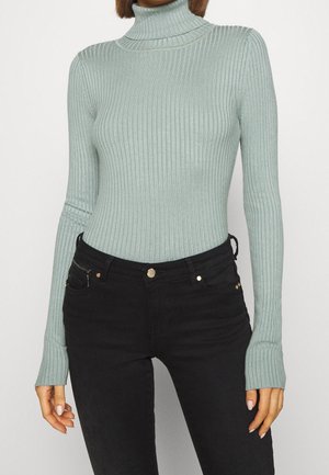 Femme portant un pull à col roulé côtelé vert clair et un jean noir taille haute, debout devant un fond uni.