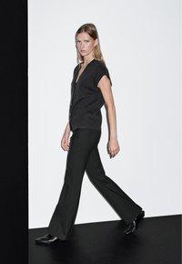 Frau, die seitwärts geht und eine dunkle ärmellose Strickjacke, schwarze Schlaghosen und schwarze spitze Schuhe trägt, vor einem schwarz-weißen Hintergrund.