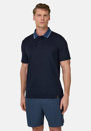 Mann trägt ein marineblaues Kurzarm-Poloshirt mit einem blau gestreiften Kragen und strukturierte marineblaue Shorts vor einfachem Hintergrund.
