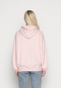 Felpa oversize rosa chiaro con cappuccio regolabile, polsini e orlo a coste, realizzata in tessuto morbido. Indossata con jeans blu chiaro.