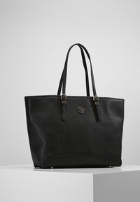 Černá kožená taška přes rameno s dvojitými ramenními popruhy, stříbrnými detaily s přezkou a malým kulatým kovovým logem na přední straně, vystavená na bílém podstavci.