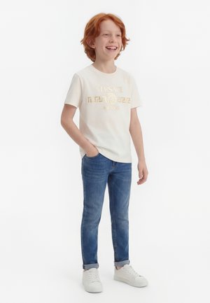 Κοκκινομάλλης αγόρι που στέκεται, χαμογελώντας, φοράει λευκό t-shirt Versace Milano, μπλε τζιν με ρεβέρ και λευκά αθλητικά παπούτσια σε λευκό φόντο.