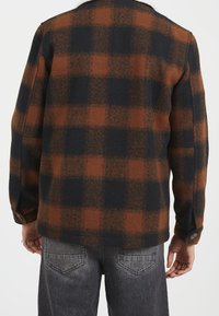 Brun och svart rutig flanelljacka med krage, knäppning vid ärmsluten och en mjuk textur, bärs över grå denim.