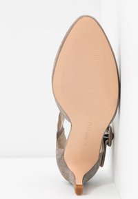 Harmaan mokkanahkakenkä, jossa on korkea korko, terävä kärki, leikattuja sivuja ja beige, teksturoitu pohja. Kengässä on pieni yksityiskohta kannassa.