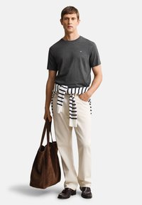 Mann trägt dunkelgraues kurzärmeliges Hemd, off-white Hosen, gestreiften Pullover um die Taille gebunden, dunkle Schuhe, hält große braune Ledertasche.