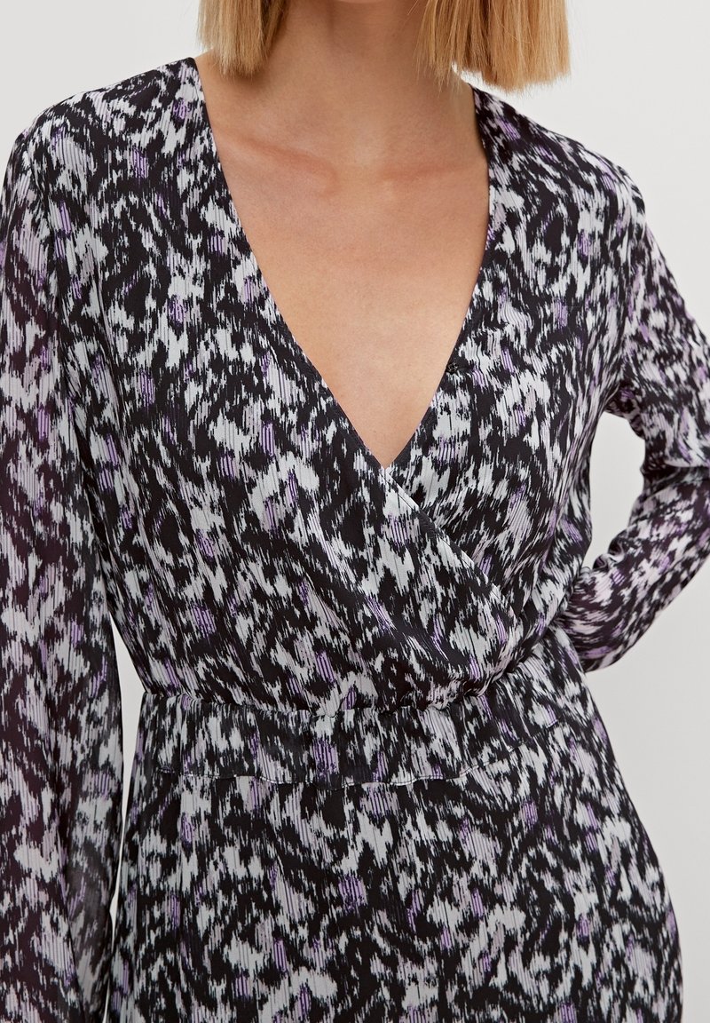 Femme portant une robe portefeuille à manches longues noire, blanche et violette à motifs, avec un profond décolleté en V et une taille élastique.
