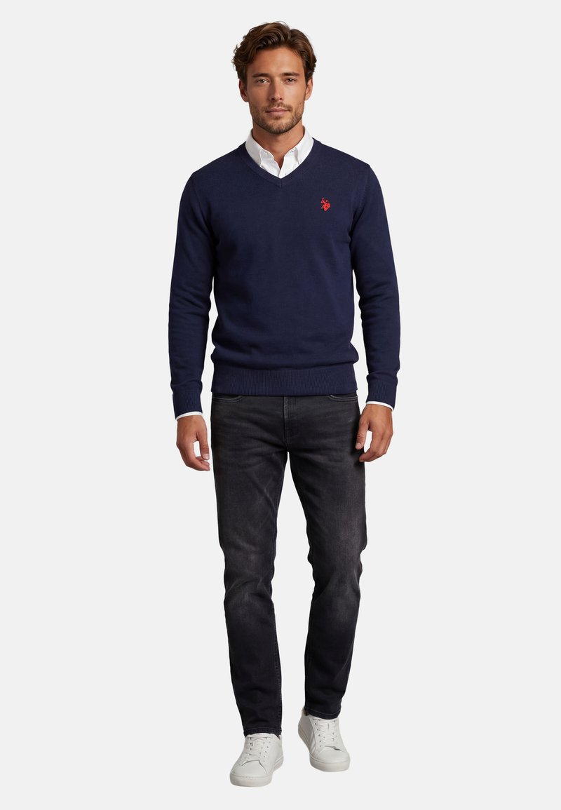 Mann trägt einen dunkelblauen V-Ausschnitt-Pullover über einem weißen Hemd, schwarze Jeans und weiße Turnschuhe, steht vor einem einfarbigen Hintergrund.