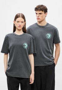 Δύο μοντέλα που φορούν γκρι βαμβακερά μπλουζάκια με στρογγυλή λαιμόκοψη και κοντά μανίκια, διακοσμημένα με ένα ανοιχτοπράσινο λογότυπο στο στήθος.