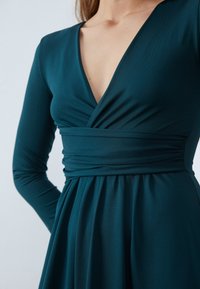 Teal klänning med långa ärmar och djup V-ringning. Tyget har en jämn yta, med en rynkad midja och flytande kjol.