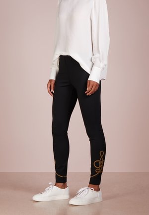 Nainen pukeutuneena istuviin mustiin housuihin, joissa on kullanvärinen kirjailu nilkkojen lähellä, yhdistettynä valkoiseen pitkähihaiseen puseroon ja valkoisiin lenkkareihin, seisoo beigen värisellä lattiolla.