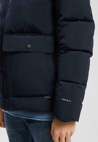 Mornarska puhovka z izdelano teksturo, stranskim žepom in zapiranjem na gumb. Vključuje logo na rokavu in pleteno plast pod njo.