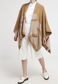 Bruine en beige gebreide poncho met open voorkant, met diagonale strepen en een zachte textuur, gedragen over witte broeken en schoenen.