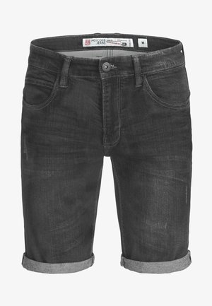 Hombre con pantalones cortos vaqueros gris oscuro remangados y zapatillas blancas, de pie frente a un fondo liso.