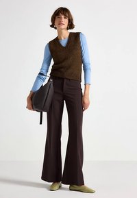 Vestă maro tricotată peste o cămașă cu mânecă lungă albastru deschis, asortată cu pantaloni evazați de culoare închisă. Modelul ține o geantă neagră texturată, purtând balerini verzi.