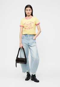 Γυναίκα που φοράει κίτρινο μπλουζάκι με σχέδιο, ανοιχτό μπλε τζιν παντελόνι με φαρδιά πόδια και ξεφτισμένα τελειώματα, μαύρα μποτάκια αστραγάλου, κρατώντας μαύρο κυλινδρικό τσάντα.