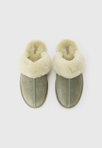 Een paar groene suède instappantoffels met een dikke crème-kleurige wolvoering, zij aan zij gerangschikt op een witte achtergrond.