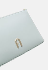 Světle modrá kožená kabelka na ruku s hladkou texturou, která má zlatý kovový doplněk ve tvaru písmene A a zipový uzávěr podél horní hrany.