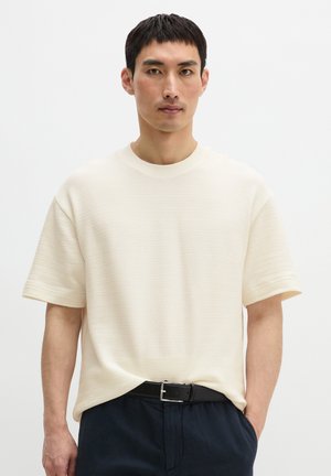 Mann trägt einen strukturierten kurzärmeligen Off-White-Pullover, in dunkle Hose mit schwarzem Gürtel gesteckt, steht vor einfachem weißem Hintergrund.
