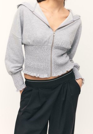 Femme portant un sweat à capuche court gris clair zippé avec taille et manches smockées, associé à un pantalon noir plissé taille haute.